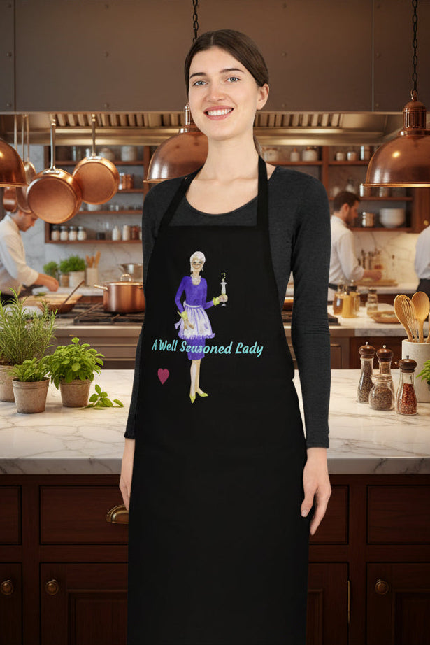 Chef wearing a black apron with an illustration of 'A Well Seasoned Lady' and 100% Cotton