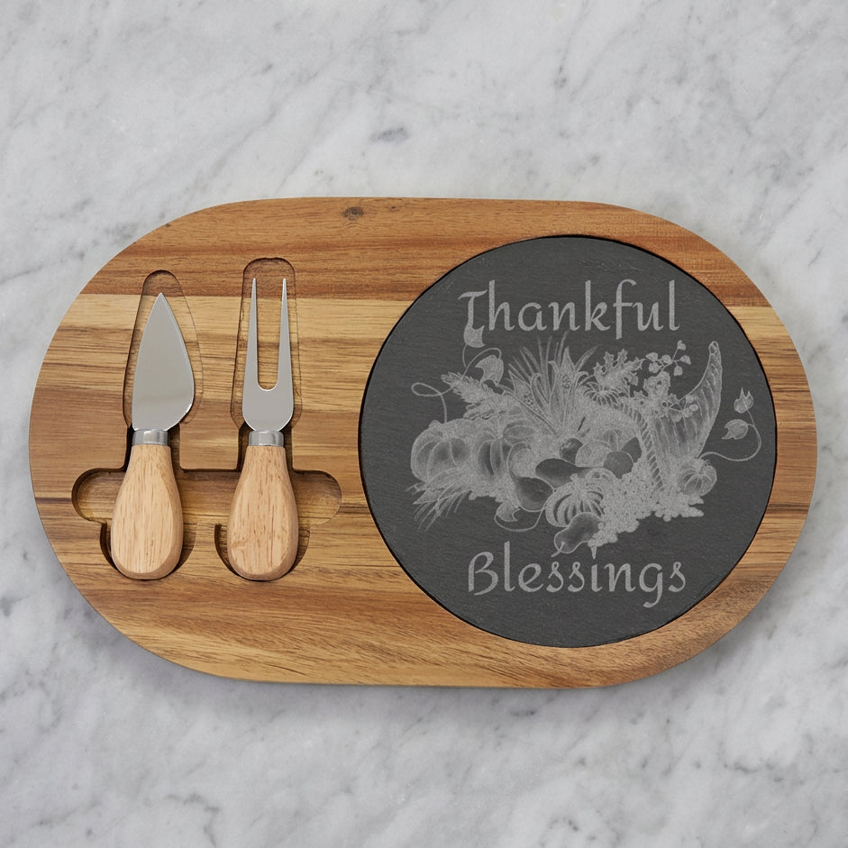 An oval wooden charcuterie board with two utensils in cutouts and a round dark slate insert engraved with artwork and the words 'Thankful Blessings'