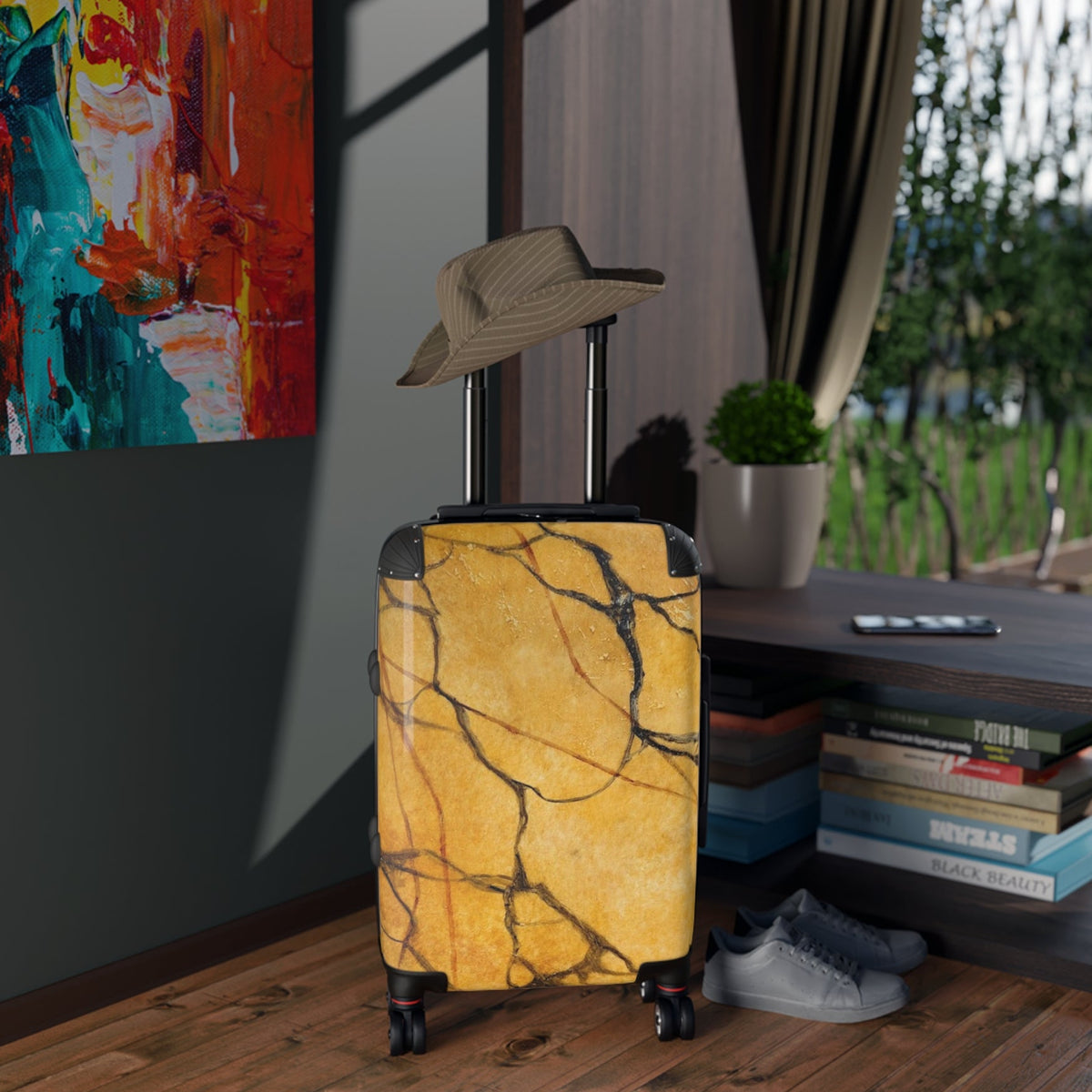Gold marble-patterned suitcase with a hat on top, placed indoors near a window with a view of greenery.