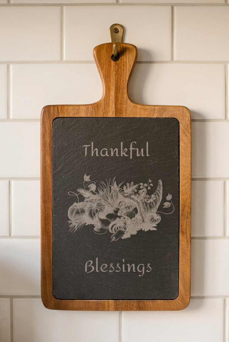 A rectangular wooden serving board with a hinged handle, featuring a dark slate insert engraved with the words 'Thankful Blessings' and a cornucopia illustration.