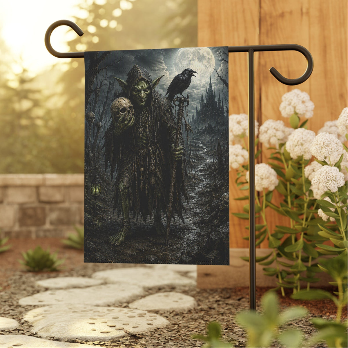 A vertical garden flag with a gothic goblin illustration hanging from a black metal stand in a garden setting. The flag depicts a dark, eerie forest scene with a hooded figure, a skull, and a raven under a full moon.