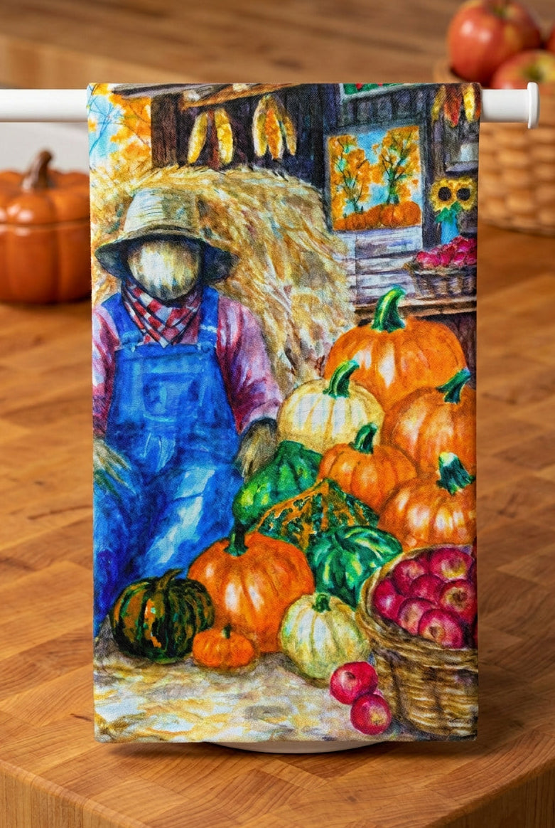 A Tea Towel art print depicting a colorful autumn harvest scene with a scarecrow, pumpkins, gourds, apples, and hay
