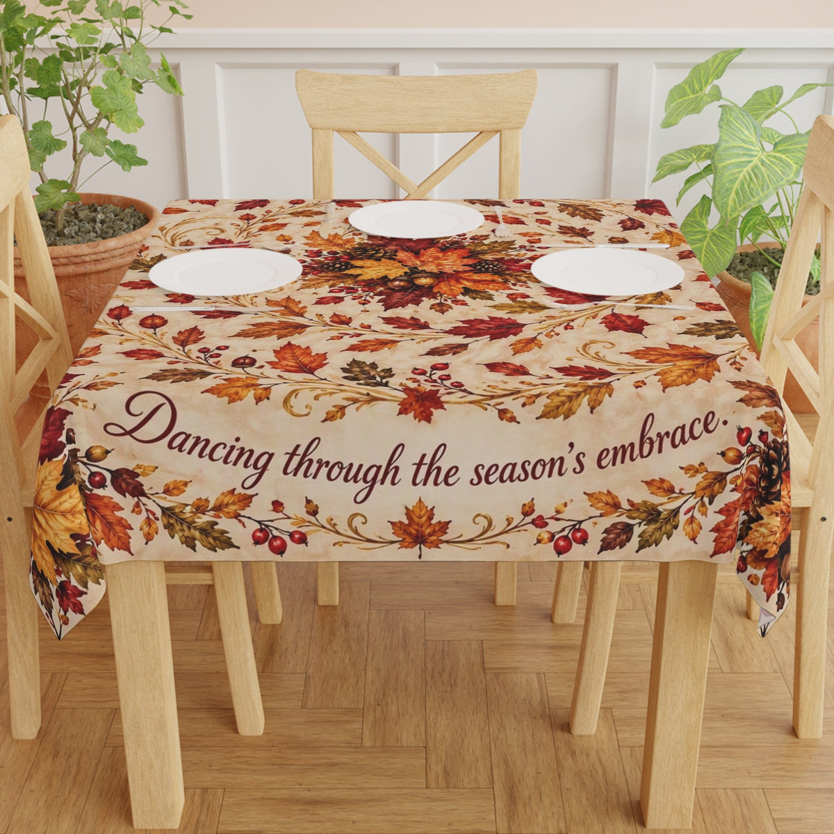 A rectangular table covered with a beige autumn-themed tablecloth featuring a floral and leaf pattern in red, orange, yellow, and brown, with the phrase 'Dancing through the season's embrace' printed across the front.