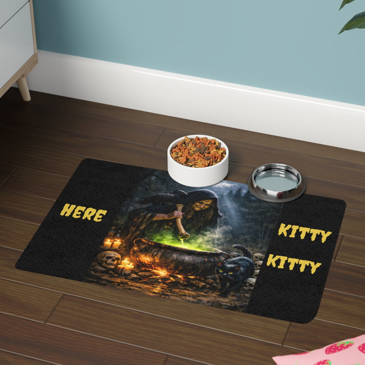 A rectangular pet mat with a dark witchy cauldron design. The mat has a black background with a glowing green and orange magical spooky scene in the center, yellow text reading 'HERE' on the left and 'KITTY KITTY' on the right