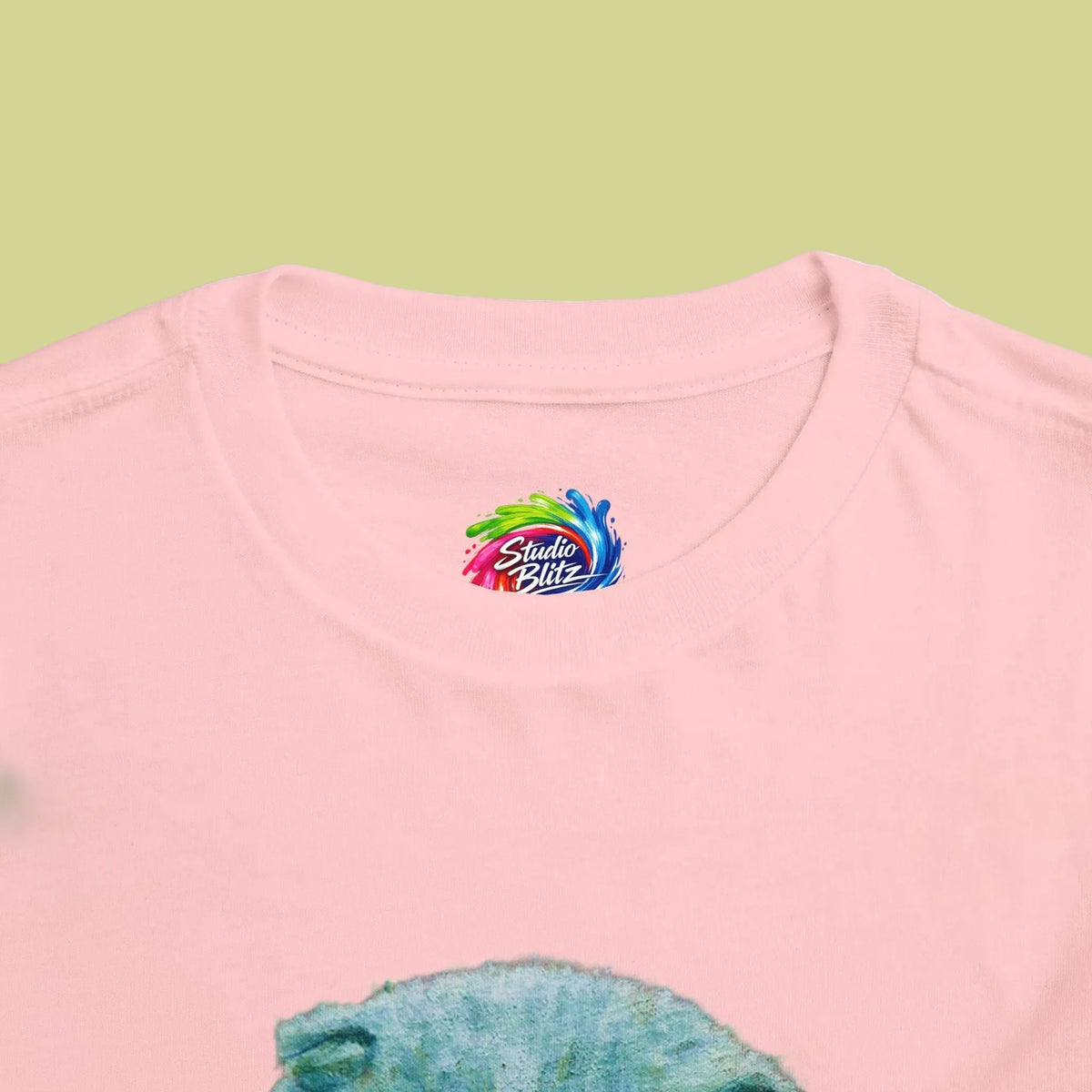 Close-up of a light pink crewneck t-shirt featuring a colorful 'Studio Blitz' logo printed inside the collar, with a blue textured graphic partially visible at the bottom.